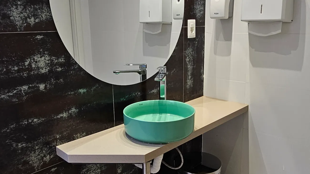 Baño de Espacio 1010 con bacha redonda color verde agua sobre mesada clara, espejo ovalado y revestimiento oscuro