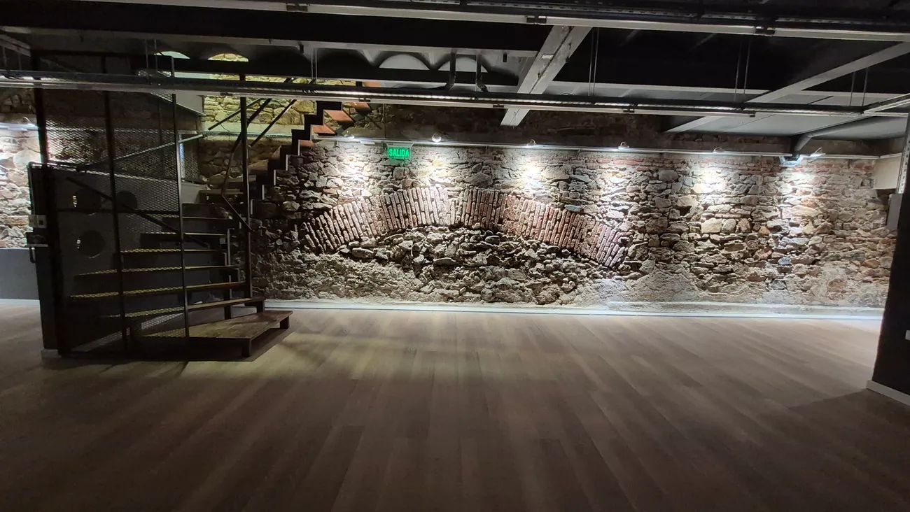 Sala amplia del subsuelo de Espacio 1010 con muro de piedra, piso de madera y escalera metálica